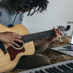 Person playing an acoustic guitar while sitting at a keyboard. Close-up view of hands on guitar strings and fretboard.