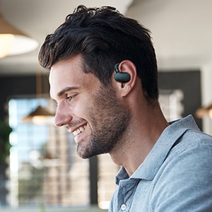 Wireless earbud worn in ear, featuring a compact dark-colored design against an office background.