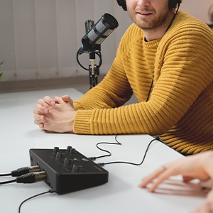Podcast setup with condenser microphone on stand, audio interface, and person wearing yellow sweater seated at table.