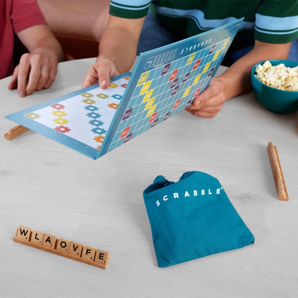 Le texte se lit comme suit : « SCRABBLE ». Plateau de jeu avec des carreaux bleus visibles, des carreaux de lettres en bois orthographiant « BIENVENUE » et un sac à cordon bleu sur la table avec un bol à pop-corn.
