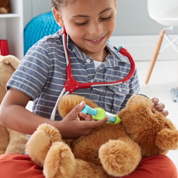 A person wearing a striped shirt and red apron examines a plush brown teddy bear with medical play equipment.