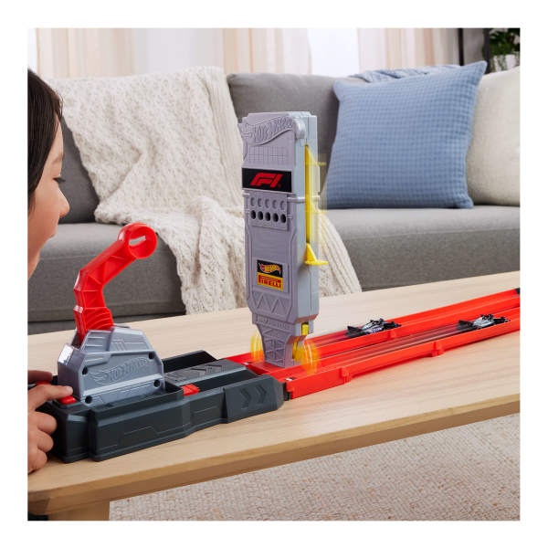 Hot Wheels-Strecken-Setup-Sequenz, in der rote Launcher und orangefarbene Streckenteile auf dem Boden in der Nähe einer grauen Couch zusammengebaut werden.